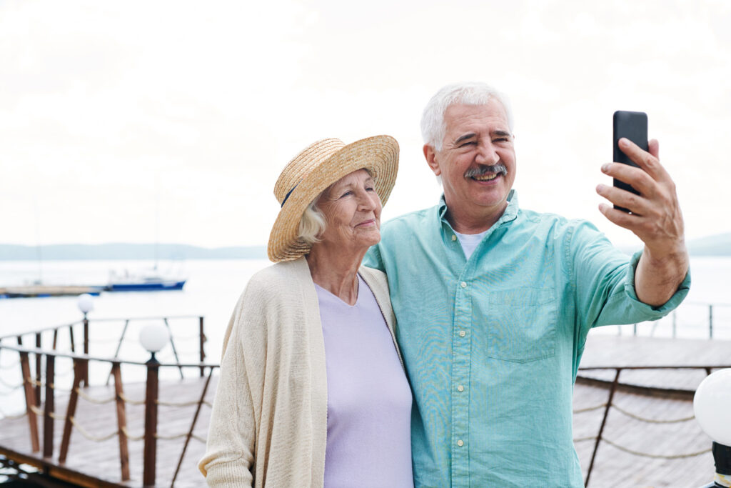 Cheerful senior husband and wife making selfie on smartphone on background of waterside on summer day